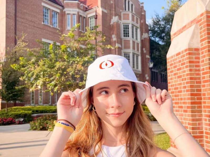 Ufora Student Living Apartments Young woman adjusts white hat, standing under "University of Florida" arch on a sunny campus day.