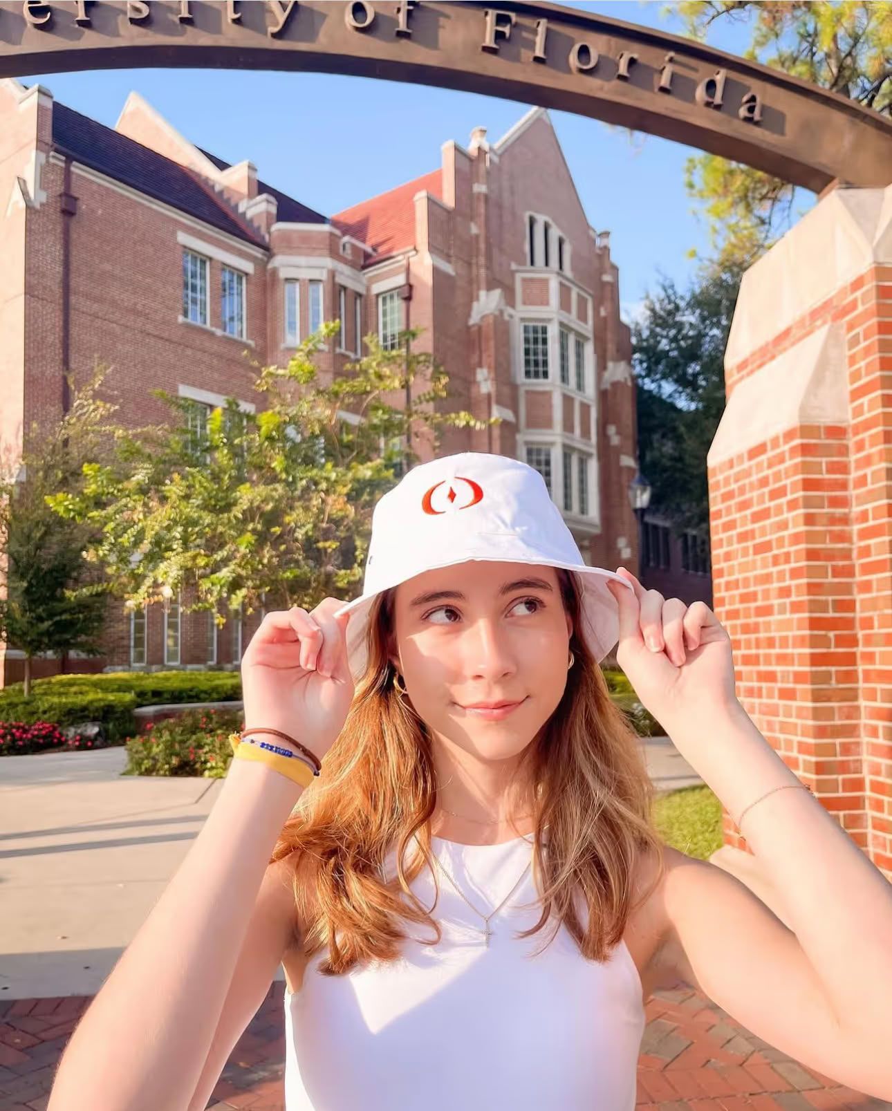 Ufora Student Living Apartments Young woman adjusts white hat, standing under "University of Florida" arch on a sunny campus day.